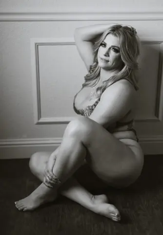 Smiling woman in lace lingerie sitting on a floor in a moody, black and white indoor portrait.