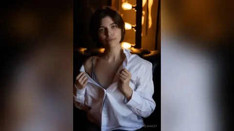 Young woman in a white button-down shirt and grey camisole posing in moody, warm indoor lighting.