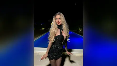 DaisyLin poses in a black lace dress with long blonde hair, standing next to a lit swimming pool at night.