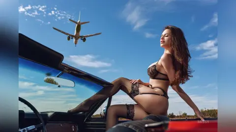 Glamorous woman in black lace lingerie relaxing on a vintage car under a bright blue sky with a plane flying above.
