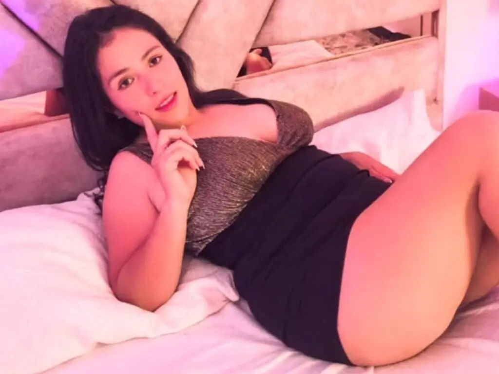 Young woman in a black and gold dress relaxing on a white bed in a soft pink lit bedroom.