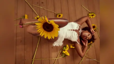Woman in white summer dress lying on wooden deck surrounded by bright sunflowers in a peaceful mood.