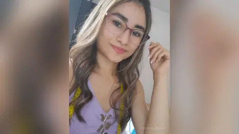 CrystallRosse wearing a purple top and glasses, smiling at the camera with her hand near her face.