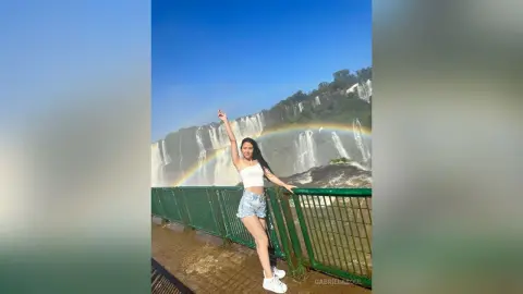 Happy woman in white crop top and denim shorts posing by a massive waterfall under a bright blue sky.