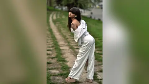 Stylish woman in white linen trousers and off-the-shoulder top posing on a park path.