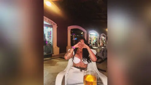 RossiNoleen wears a white lace bra and white bottoms while sitting outdoors, making a heart shape with her hands over her face.