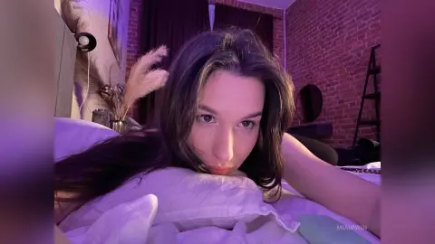 Young woman with long dark hair relaxing on a white pillow in a moody, purple-lit bedroom setting.