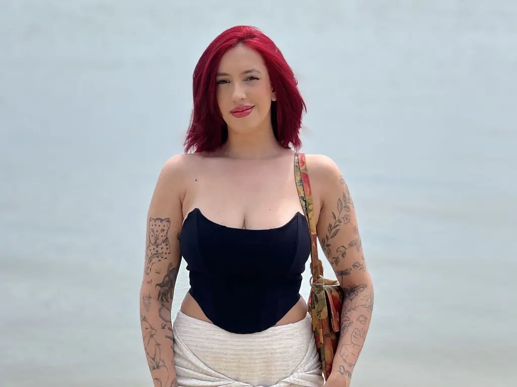 Red-haired woman in a black corset top and white skirt standing by the ocean on a sunny day.