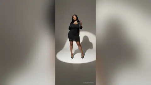Elegant woman in a chic black cocktail dress posing confidently against a minimalist grey studio background.