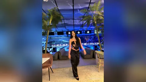 Cheerful woman in a black bodysuit and trousers standing in a modern tropical-themed lounge with palm trees.