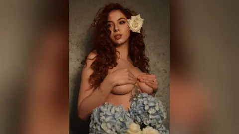 Redheaded woman in a floral blue hydrangea outfit posing with a white rose in a moody, artistic setting.