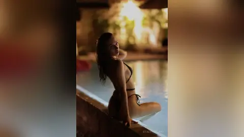 StacyHarland poses in a black bikini while sitting on the edge of a swimming pool at sunset.