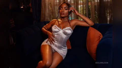 Glamorous woman in a white satin lace bodysuit relaxing on a navy velvet sofa in moody indoor lighting.