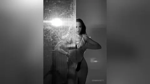 Moody black and white portrait of a woman in elegant lingerie posing behind a textured glass partition.