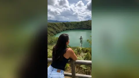 FabianaLima looks out over a green lake while leaning against a wooden fence in a black tank top and light blue denim jeans.
