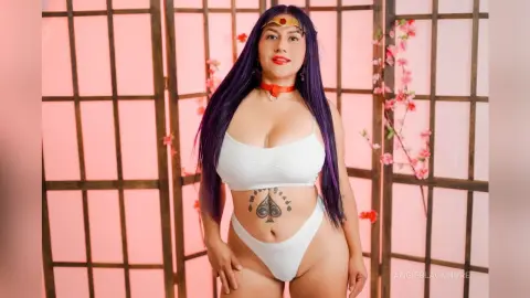 Woman with long purple hair in white bikini and gold headpiece posing against a pink floral background.