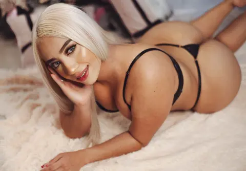 Blonde woman in black lingerie lying on a white faux fur rug in a cozy, intimate setting.