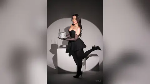 Elegant woman in a black cocktail dress holding a grey cake in a minimalist studio setting.