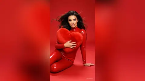 AnnaKarev poses in a tight red bodysuit while hugging a large red heart against a red background.