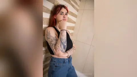 Tattooed woman in a black crop top and blue jeans posing thoughtfully against a tiled wall.