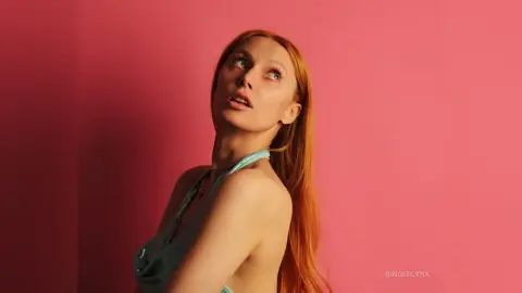 GingerLynx looks up with a pouting mouth, wearing a light blue top against a pink background.