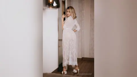 Elegant woman in a white lace maxi dress posing in a modern minimalist interior with soft lighting.