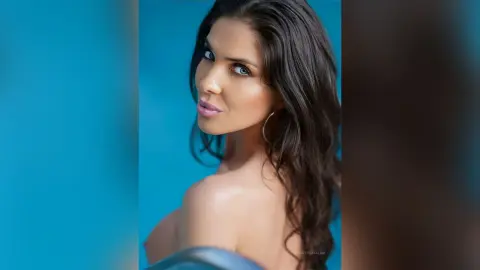 KinsleyCatalina poses with a direct gaze, showing her bare shoulders and long dark hair against a blue background.