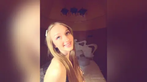Smiling young woman with long blonde hair in a cozy, dimly lit bedroom setting.