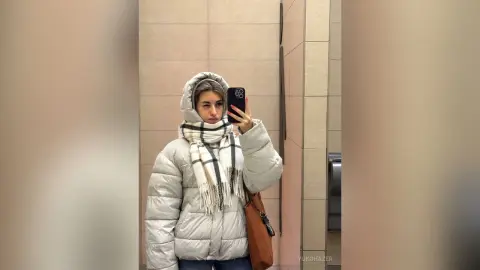 Young woman in a white puffer jacket and plaid scarf posing for a mirror selfie in a modern hallway.