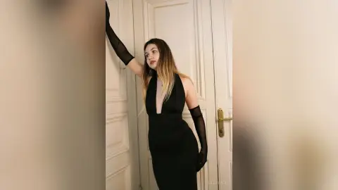 Elegant woman in a black halter evening dress and sheer gloves posing by a white door in a moody setting.