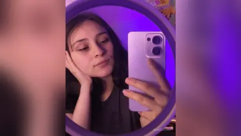 Young woman in a dark t-shirt taking a moody selfie in a purple neon lit room with a ring light.