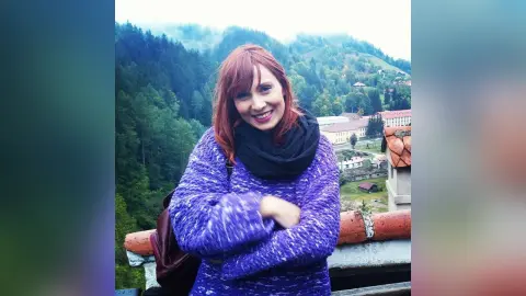 AryaSin smiles with her red hair and a black scarf, wearing a purple sweater while standing outdoors against a forest background.
