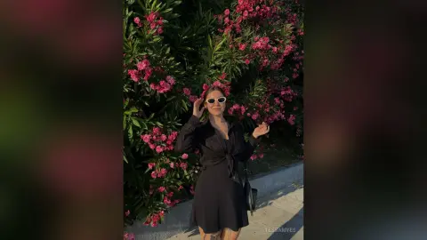 Smiling woman in a black dress and white sunglasses posing against blooming pink flowers in bright sunlight.