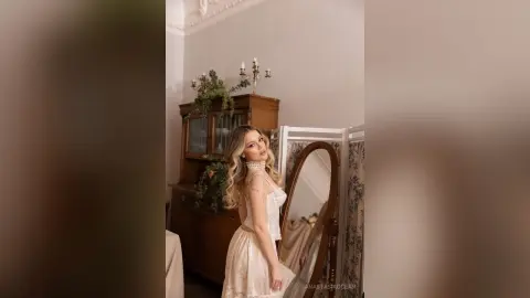 Elegant woman in a white lace dress posing in a vintage room with a classic wooden cabinet and mirror.