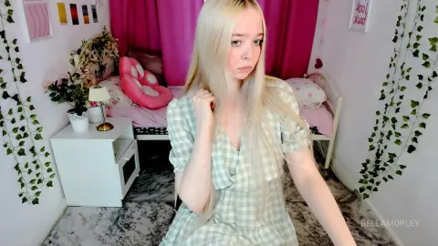 Blonde woman in a green gingham dress sitting in a cozy bedroom decorated with vines and pink accents.