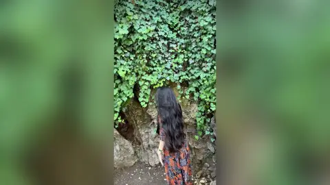 Woman in a floral patterned dress standing before a lush green ivy wall in a serene natural setting.