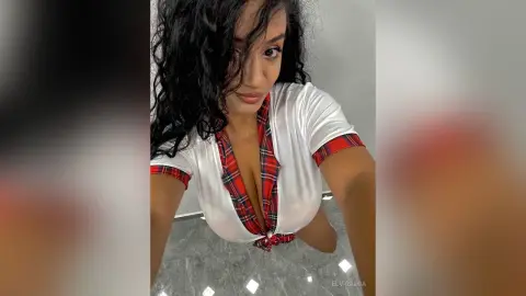 Curvy woman with long dark hair wearing a white and red plaid shirt in a bright indoor setting.