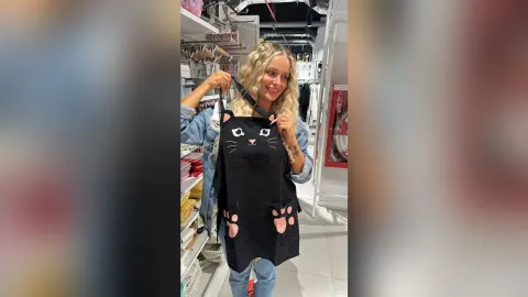 Smiling blonde woman in a denim jacket holding a black cat graphic tote bag in a clothing boutique.
