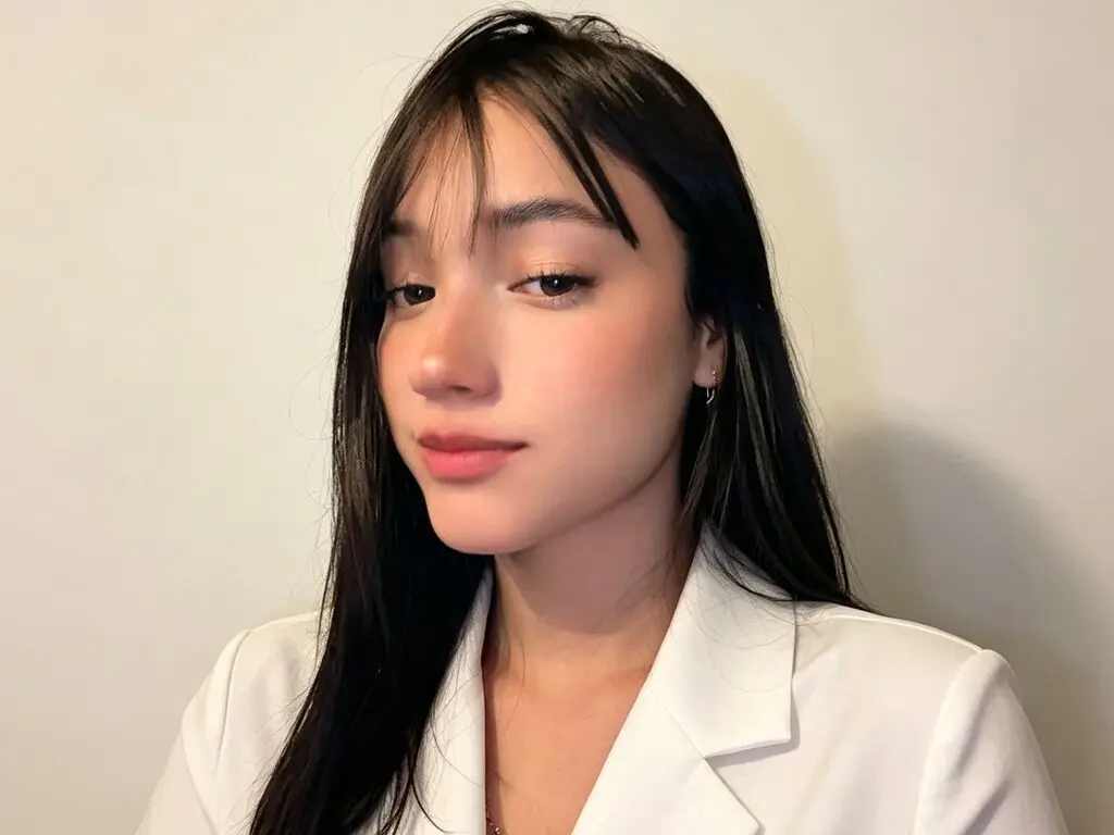 Young woman with long dark hair and bangs wearing a white blazer against a neutral background.
