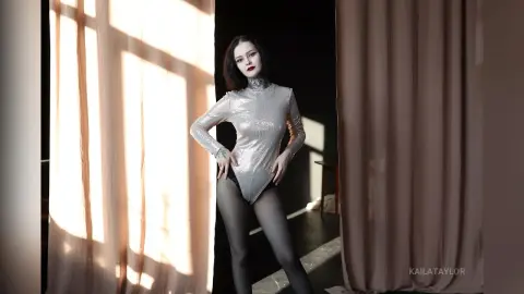 Gothic woman in a shimmering silver bodysuit and black tights posing in a sunlit studio setting.