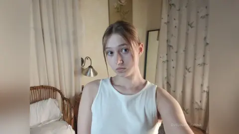Young woman in a white tank top posing in a cozy, sunlit bedroom with vintage floral decor.