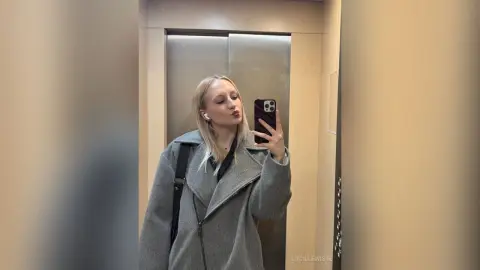Blonde woman in a grey wool coat taking a mirror selfie in a modern elevator.