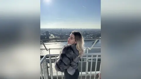 ValerieGracy poses on a balcony overlooking a city skyline wearing a thick grey fur coat.