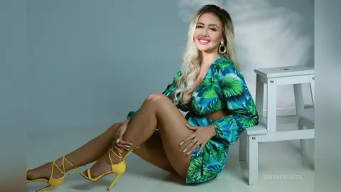 Anita sits on a white stool wearing a green tropical print wrap dress and yellow strappy heels.