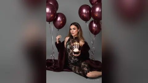 Glamorous woman in a black lace dress and faux fur coat holding a birthday cupcake with lit candles.