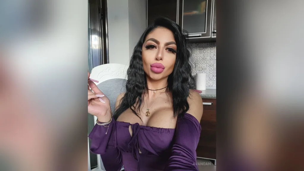 AvaNoah — AvaNoah poses in a purple low-cut top with long wavy dark hair while looking at the camera.