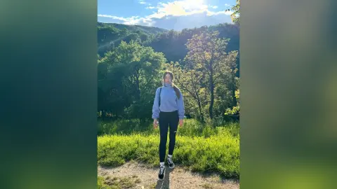 Young woman in a blue sweatshirt and black leggings standing on a forest trail under a sunny sky.
