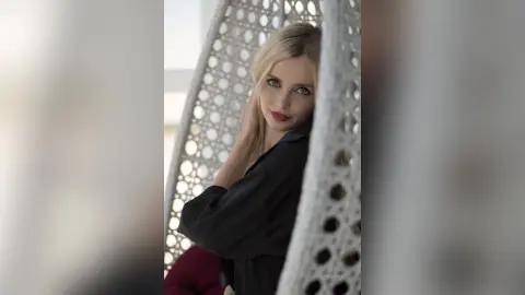 Elegant blonde woman in a black blouse relaxing in a white hanging egg chair with a serene mood.
