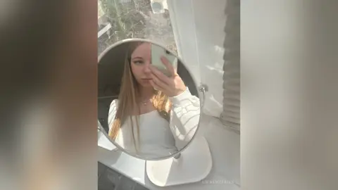 AdenaMcfalls poses for a mirror selfie wearing a white long-sleeve shirt, showing her long blonde hair and face in a bright room.