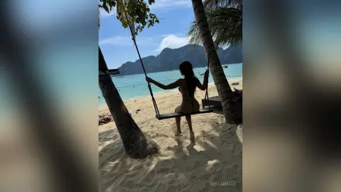 Woman in black bikini relaxing on a wooden swing under palm trees at a tropical beach resort.